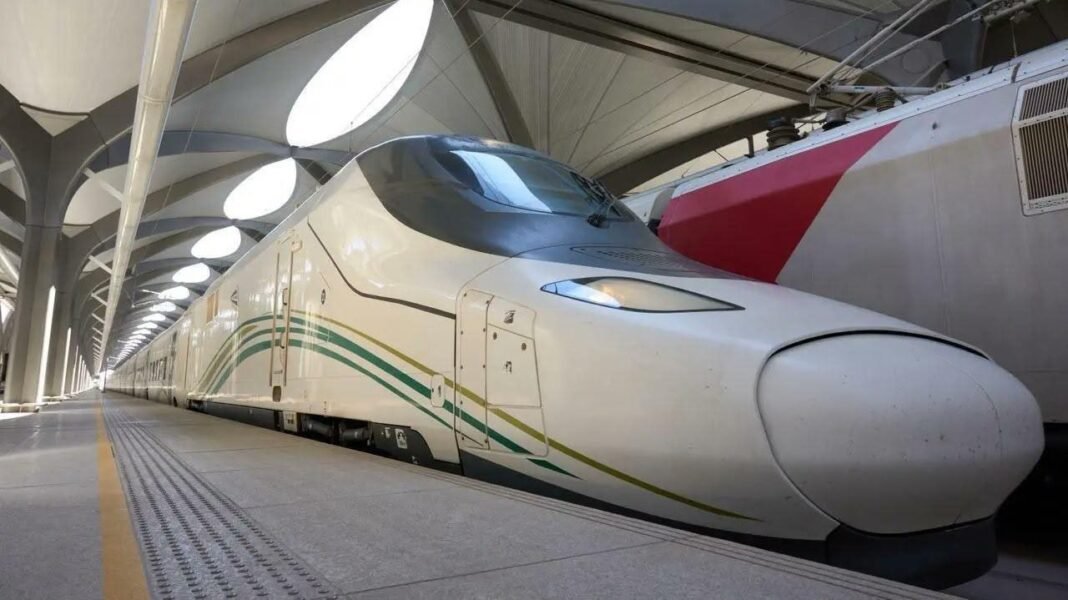 A white Haramain High Speed Railway train with green and gold stripes at a covered station platform