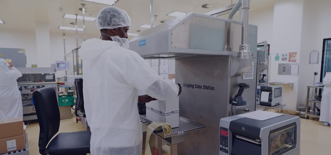 A worker in a white gown and hairnet operates packaging machinery in a bright pharmaceutical production hall