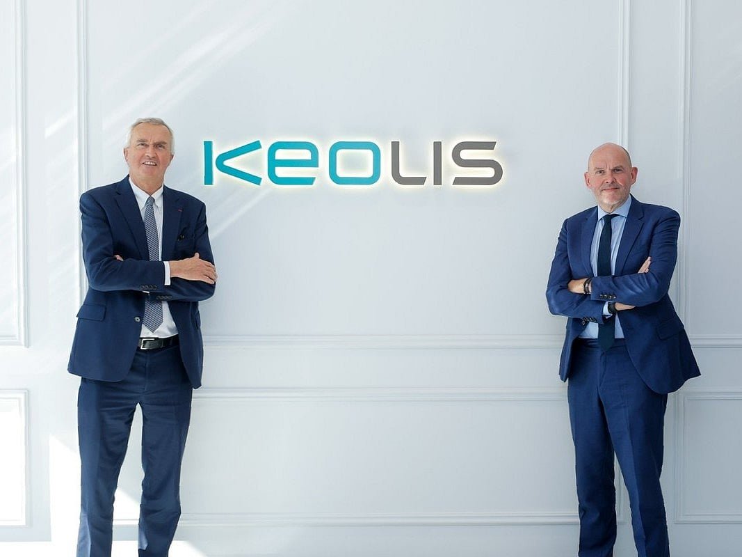 Two men in navy suits stand with arms folded on either side of an illuminated Keolis logo on a white paneled wall