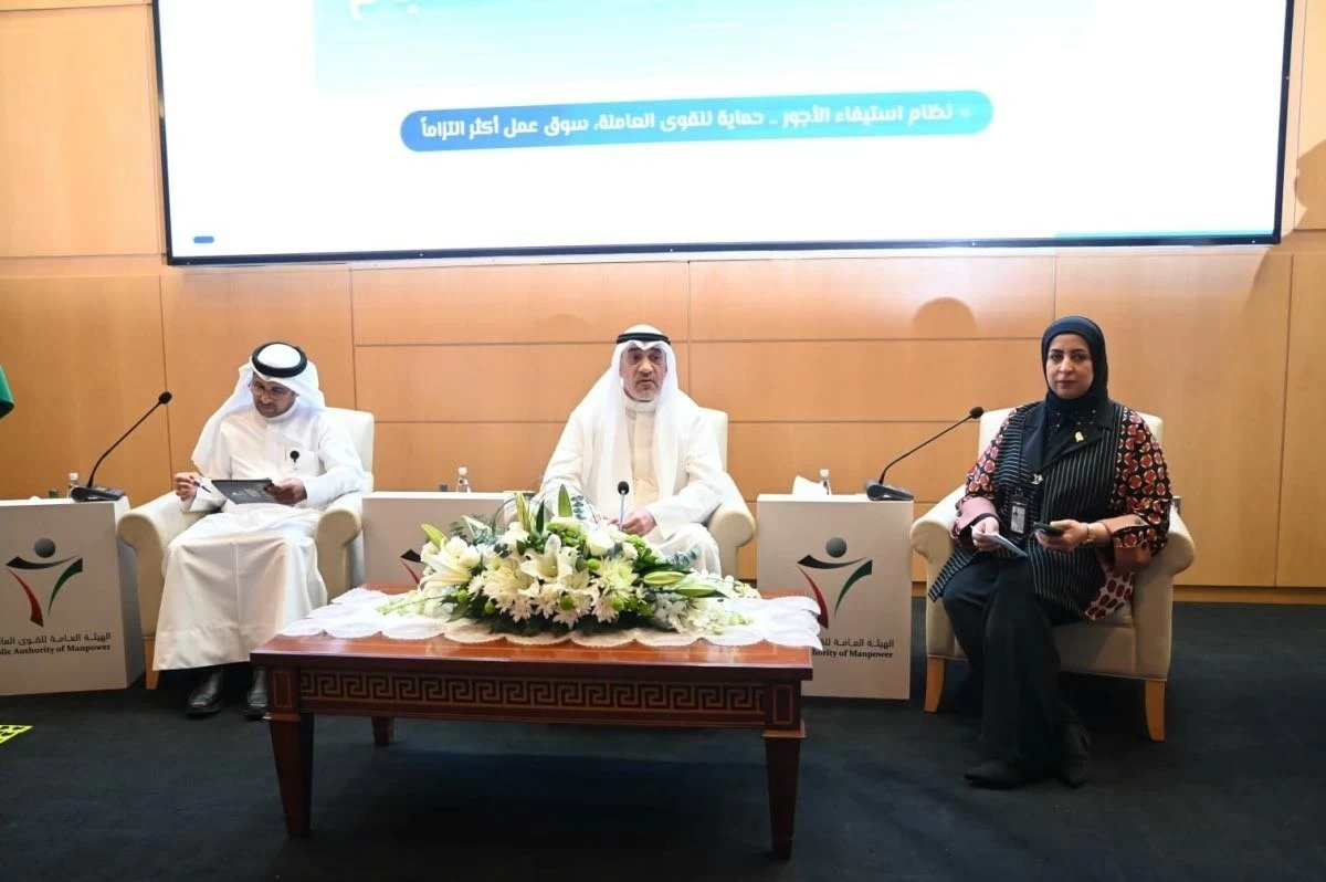 Three officials seated in armchairs on a stage behind a table of white flowers, beneath a screen with Arabic text about the wage payment system