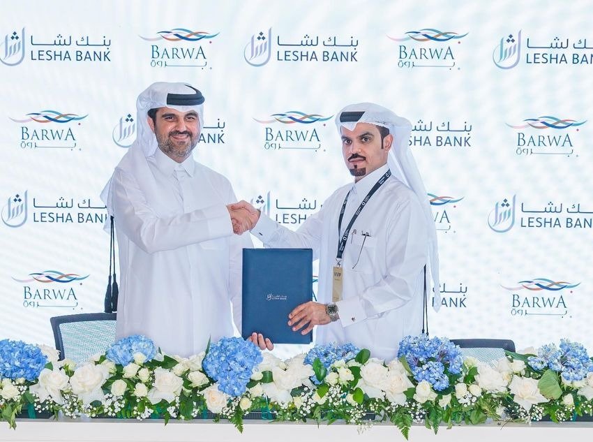 Two executives in Qatari dress shake hands and hold a blue signing folder in front of a backdrop bearing the Lesha Bank and Barwa logos