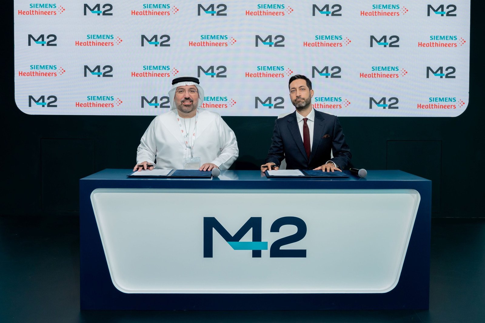 Two executives seated at a signing desk with M42 and Siemens Healthineers branding on the backdrop