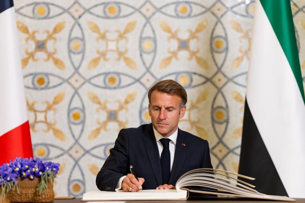 Emmanuel Macron seated at a desk signing a guestbook between French and UAE flags