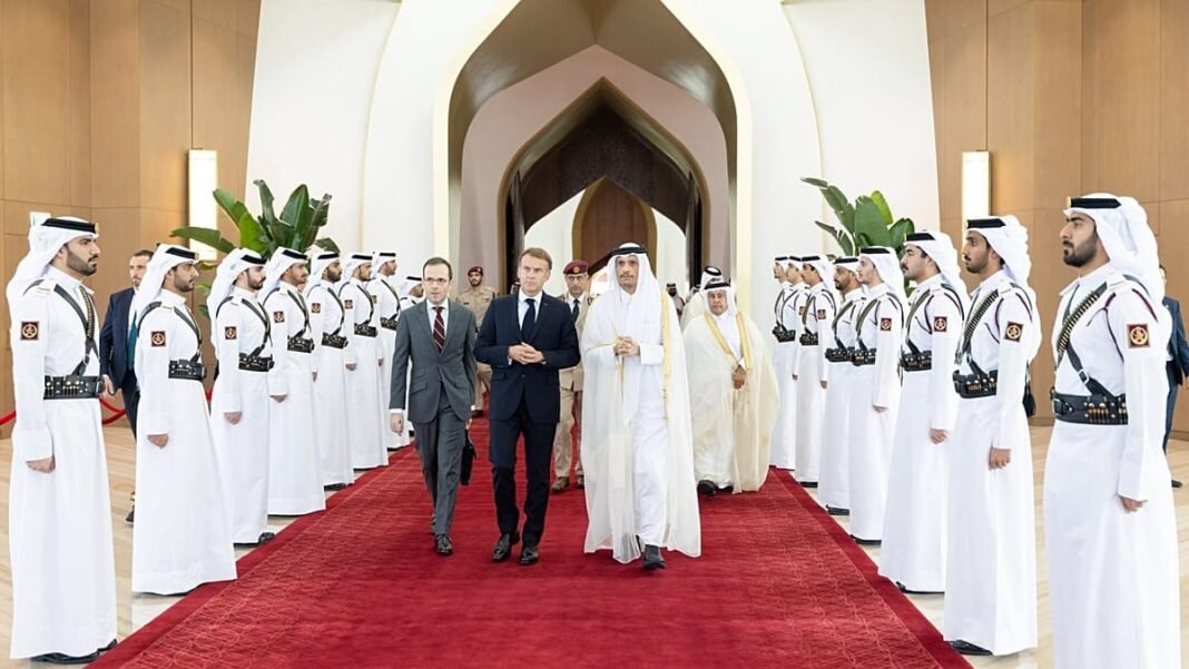 Emmanuel Macron in a dark suit walks along a red carpet beside a man in white Qatari dress, flanked by rows of honor guards in white robes
