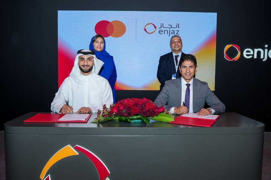 Two men sign documents at a table with red roses in front of a screen showing Mastercard and Enjaz logos while a man and a woman stand behind them