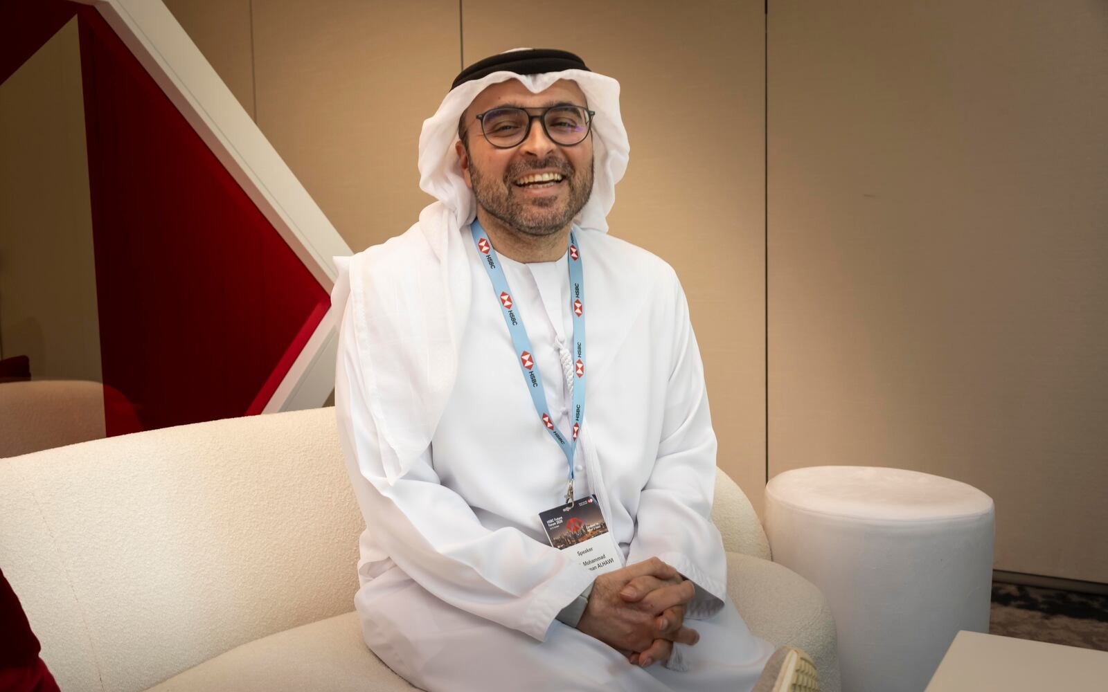 Mohammad Abdulrahman Alhawi, wearing a white kandura and an event lanyard, smiles while seated on a cream sofa