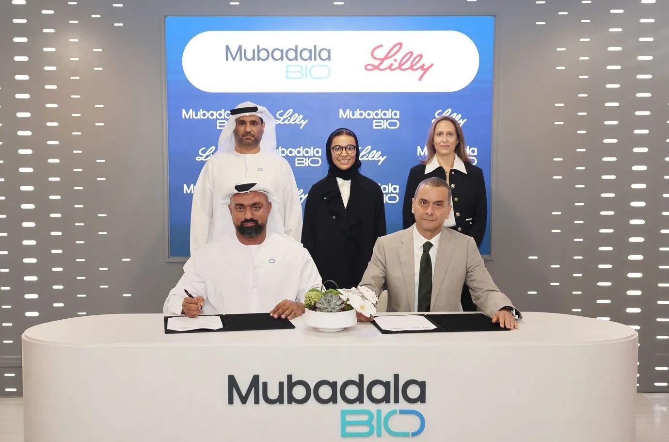 Mubadala Bio and Eli Lilly executives sign an agreement at a table in front of a Mubadala Bio and Lilly backdrop