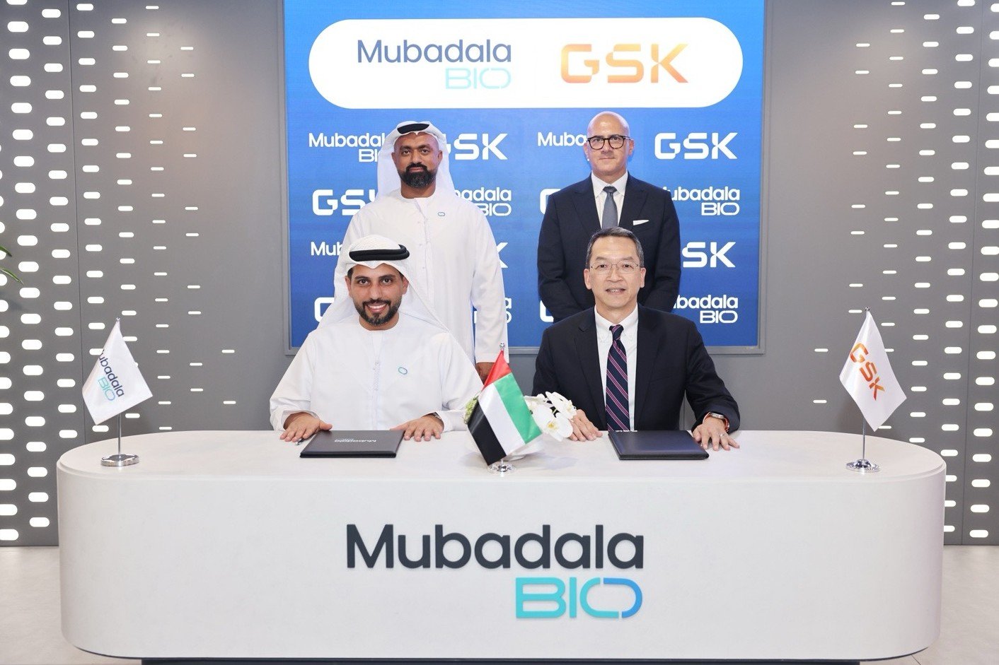 Two executives sign documents at a white Mubadala Bio desk as two colleagues stand behind them in front of a Mubadala Bio and GSK backdrop