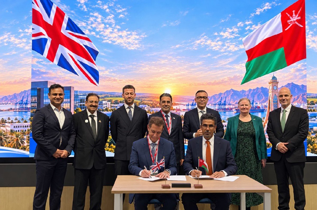 Two men sign documents at a table in front of British and Omani flags as officials and executives stand behind them