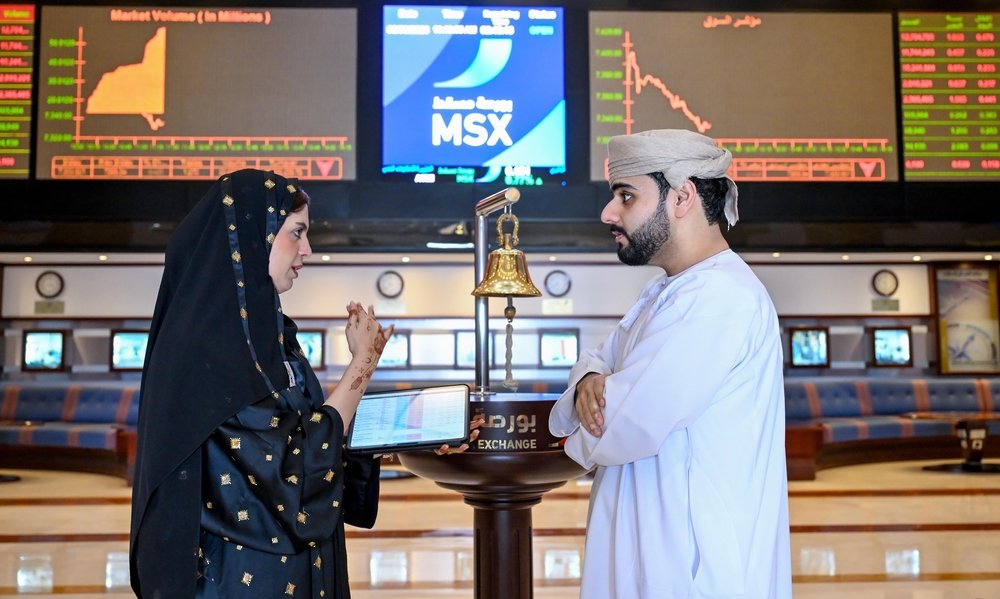A woman and a man talk beside the bell in the Muscat Stock Exchange trading hall, with market screens behind them