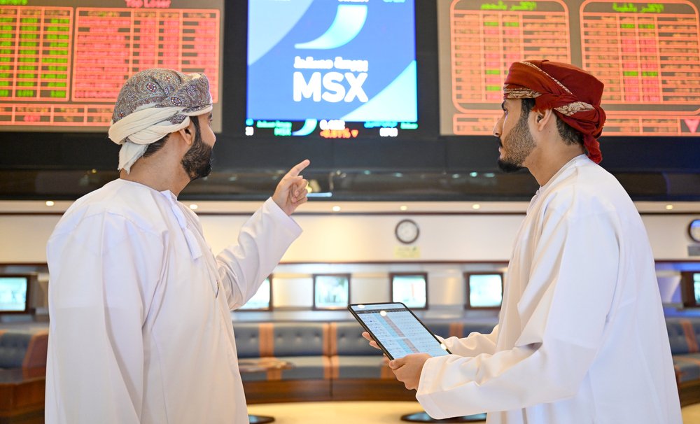 Two men in Omani dress look at trading screens at the Muscat Stock Exchange, one holding a tablet