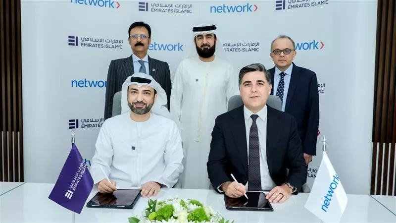 Two executives seated at a table signing an agreement on tablets, with three colleagues standing behind them in front of Emirates Islamic and Network branding
