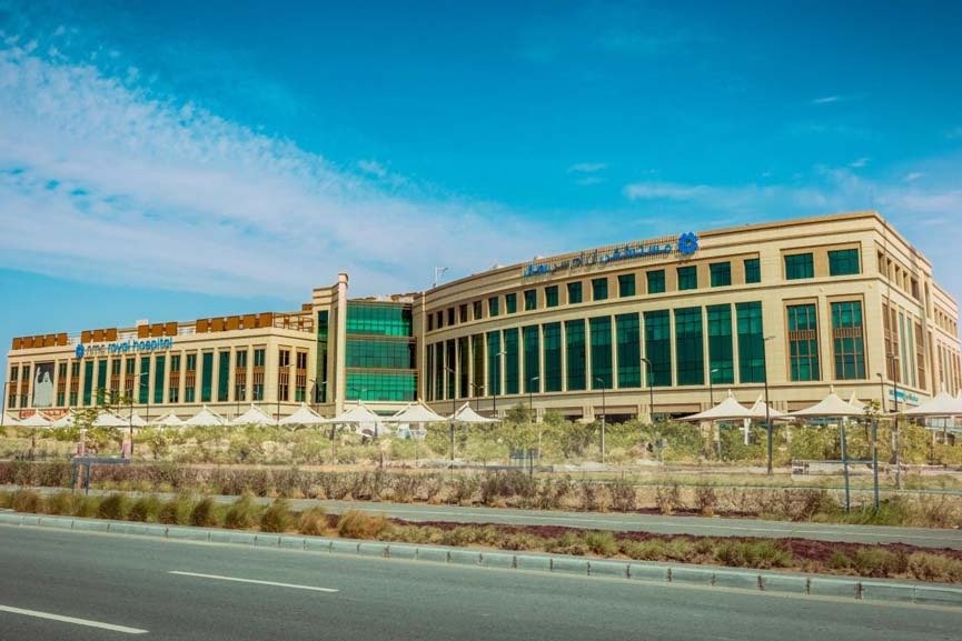 A large beige and green-glass NMC Royal Hospital building beside a road in Abu Dhabi under a blue sky