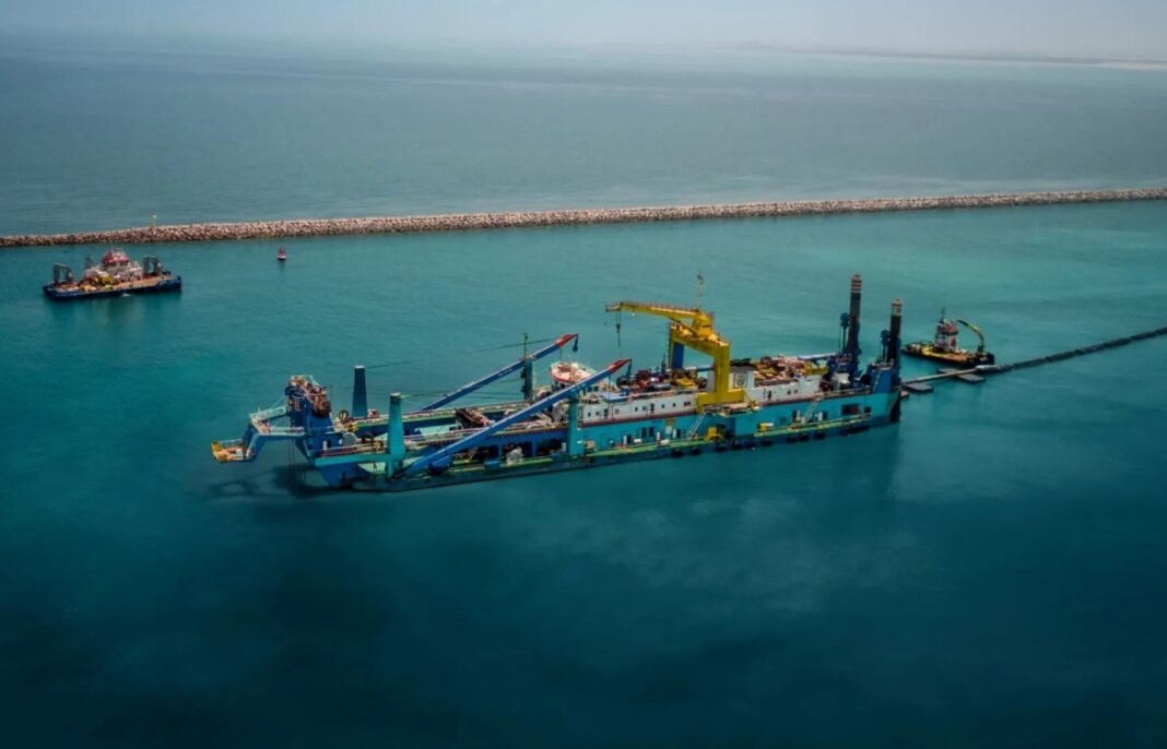 An NMDC cutter suction dredger works in turquoise water beside a rock breakwater, with support vessels nearby