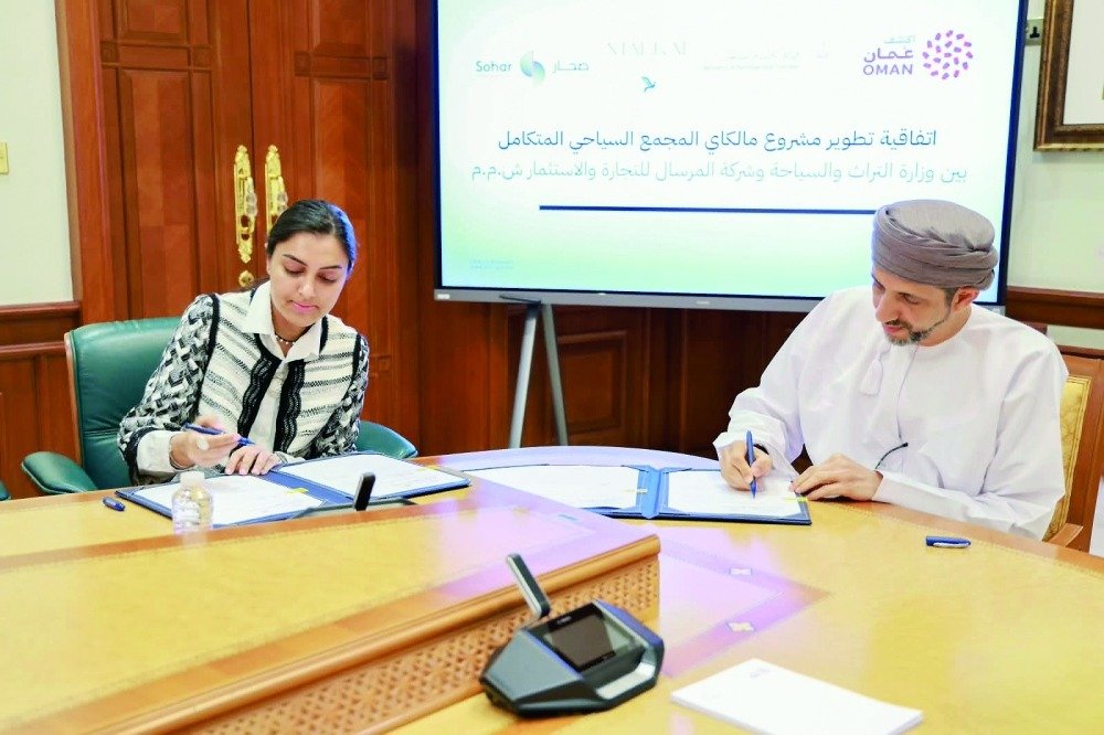 Oman's heritage and tourism minister and a businesswoman signing documents at a conference table in front of a screen announcing the Malkai agreement