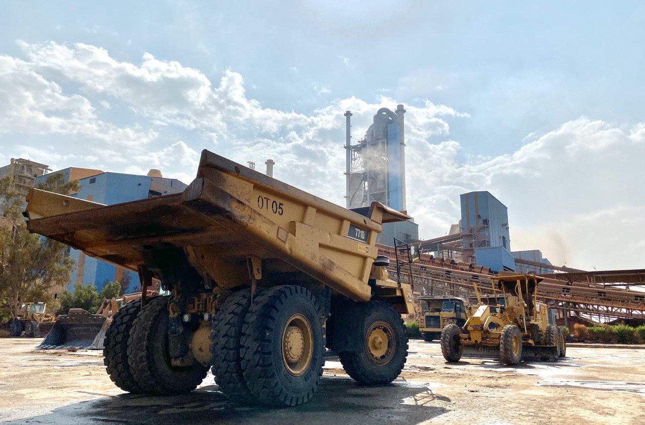 A yellow mining haul truck parked in front of a cement plant tower and conveyor belts under a cloudy sky