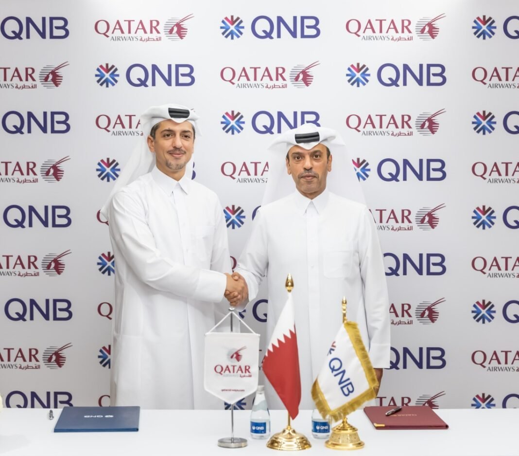 Two executives in Qatari dress shake hands behind a signing table with Qatar Airways and QNB flags