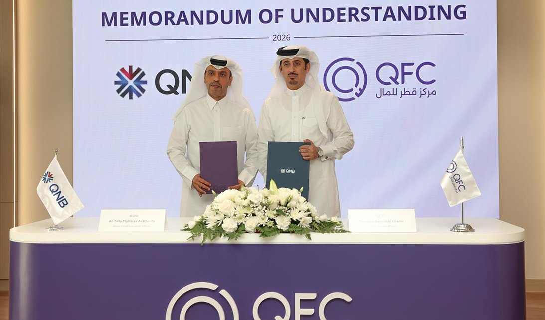 Two men in white Qatari dress hold signed folders behind a table with white flowers, in front of a screen reading Memorandum of Understanding with QNB and QFC logos