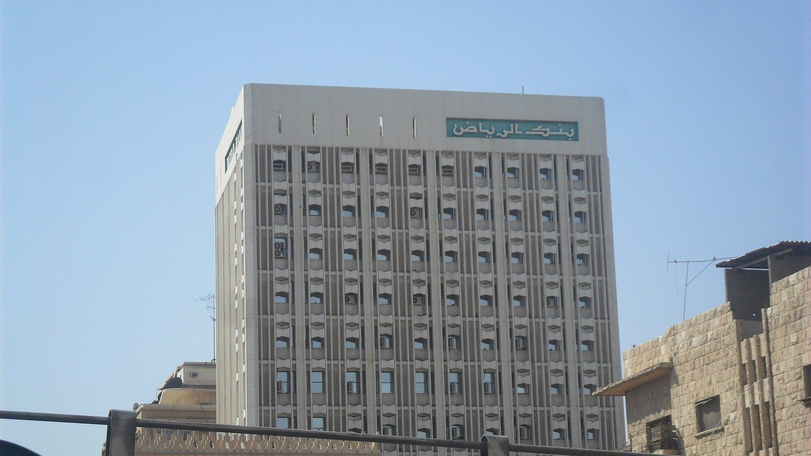 A tall pale concrete office tower with green Arabic lettering reading Riyad Bank near its roof, under a clear blue sky in Riyadh