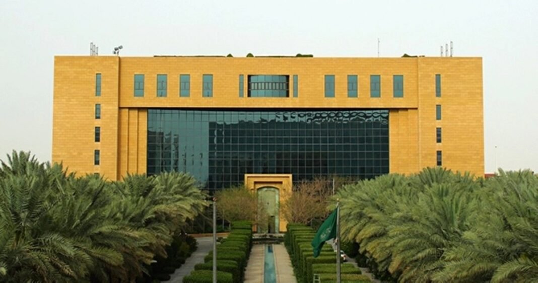 A sand-colored government building with a glass facade behind rows of palm trees and a Saudi flag