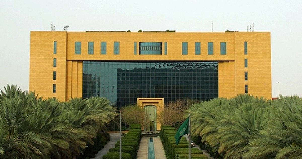 A sand-colored government building with a glass facade behind rows of palm trees and a Saudi flag