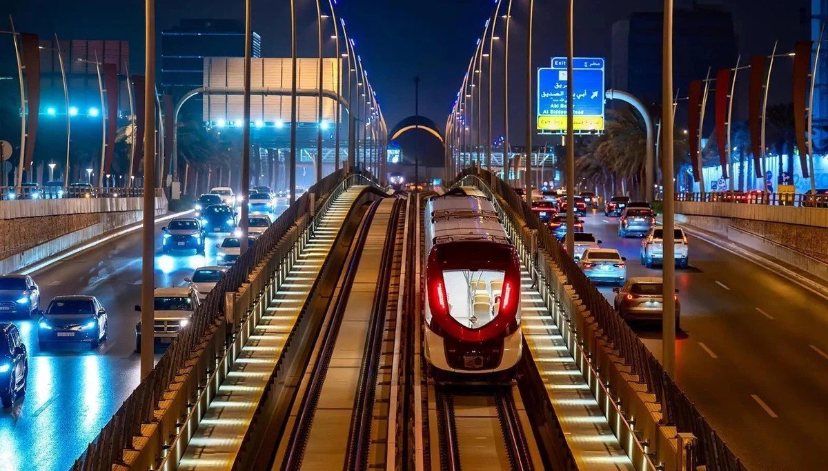 A red and white Riyadh Metro train on an elevated track at night between busy lanes of car traffic