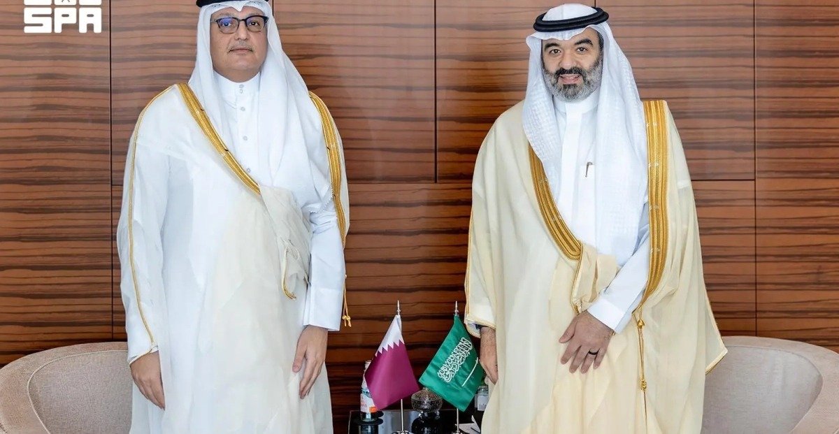 Two ministers in traditional Gulf attire stand beside desk flags of Qatar and Saudi Arabia in a wood-paneled meeting room