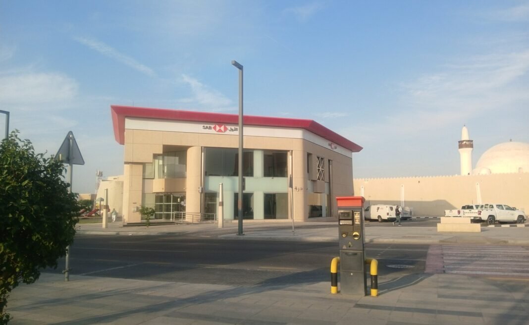 A two-story beige Saudi Awwal Bank branch building with a red roof edge and SAB sign, beside a road and a mosque with a white dome