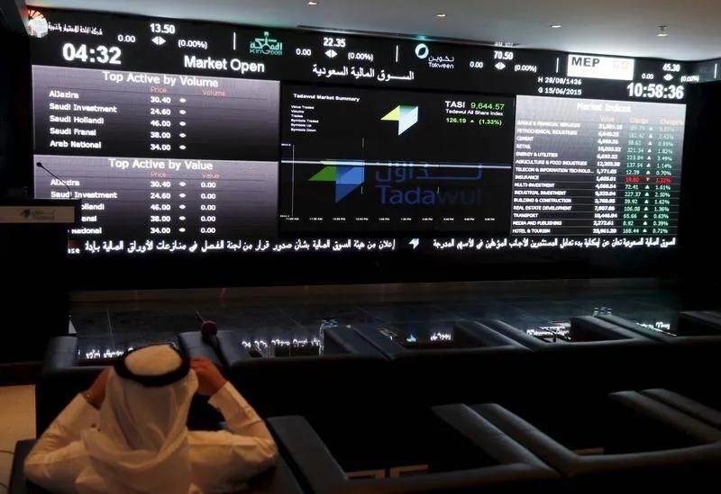 A man in a white headdress sits facing a large electronic board showing share prices and the Tadawul All Share Index at the Saudi Exchange