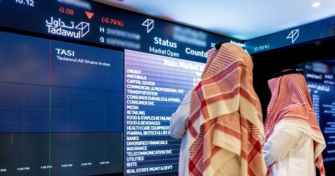 Two men in red checkered headscarves look at large trading screens showing the Tadawul All Share Index and market sectors