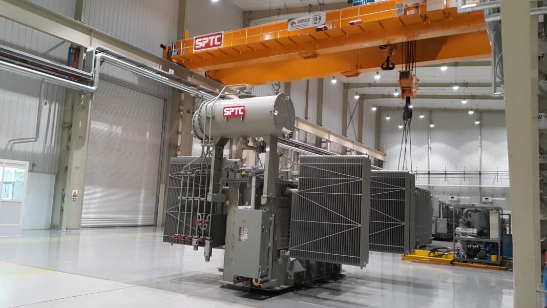 A large power transformer fitted with radiator banks inside a factory hall under an overhead crane