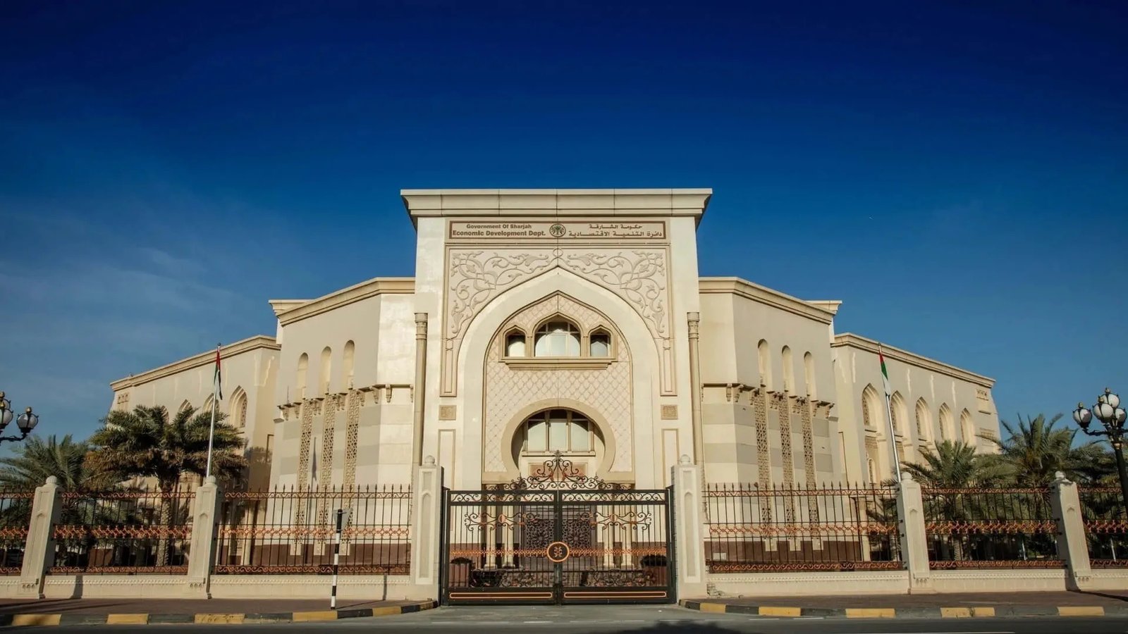 The cream-colored Sharjah Economic Development Department building with an arched entrance and iron gates under a blue sky