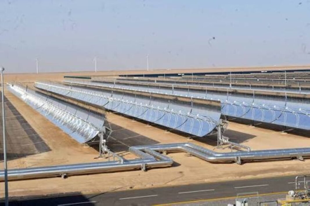 Rows of parabolic-trough solar collectors and pipes in the desert, with wind turbines in the distance