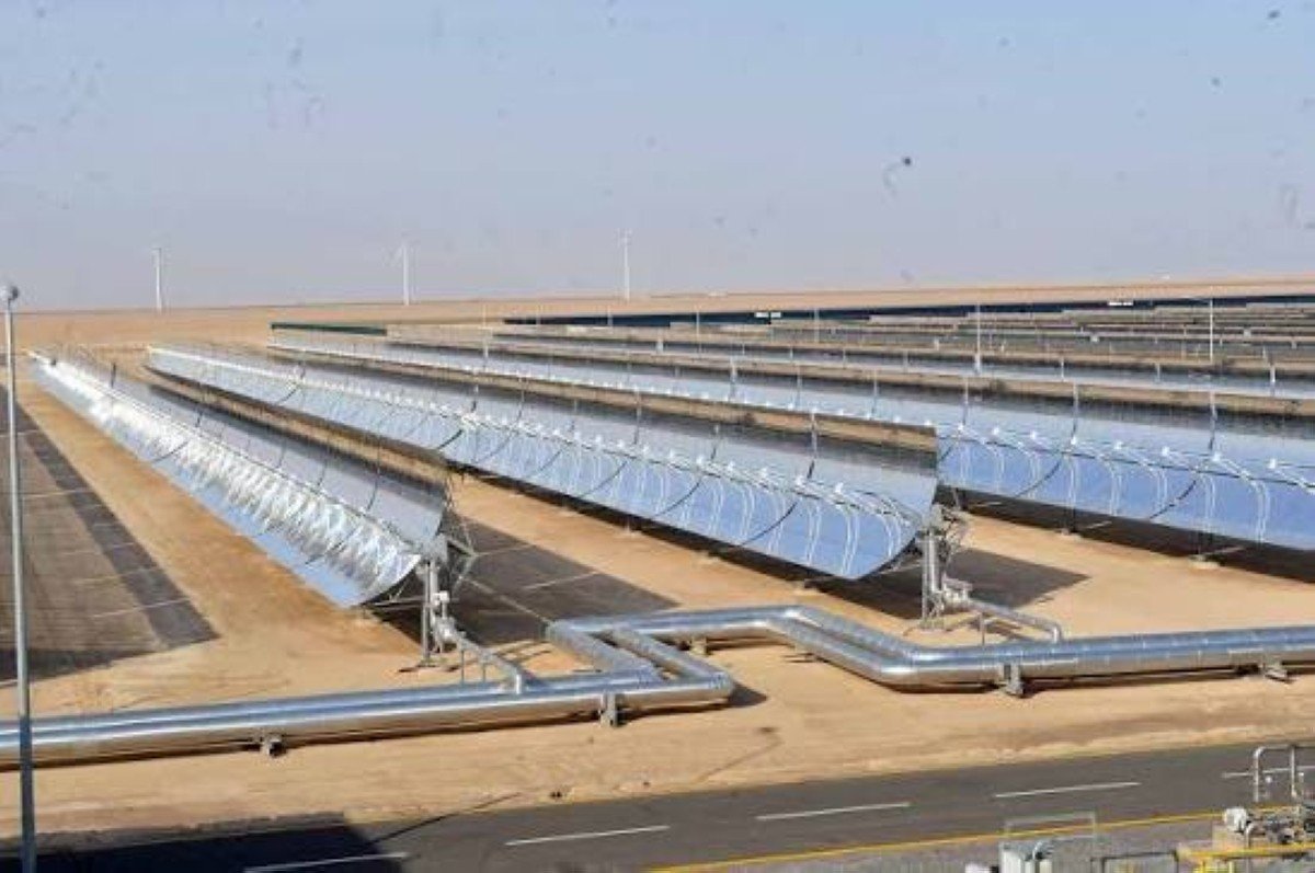 Rows of parabolic-trough solar collectors and pipes in the desert, with wind turbines in the distance