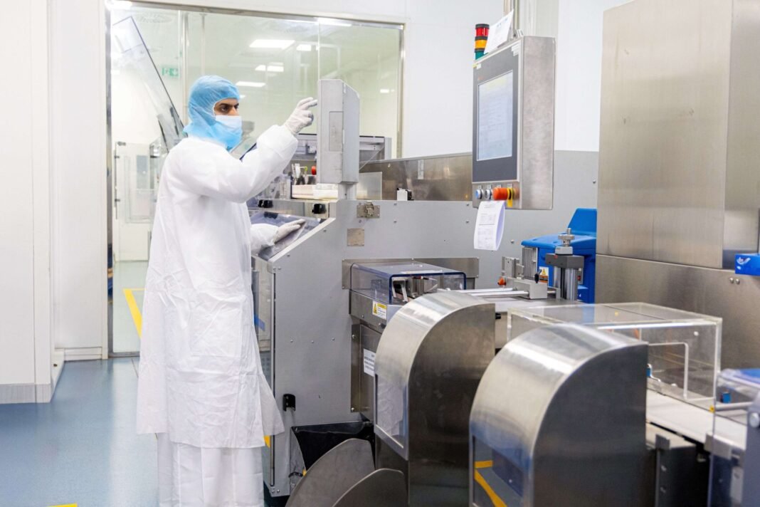 A worker in a white cleanroom suit, hairnet and mask operates a stainless steel packaging machine at a SPIMACO pharmaceutical plant