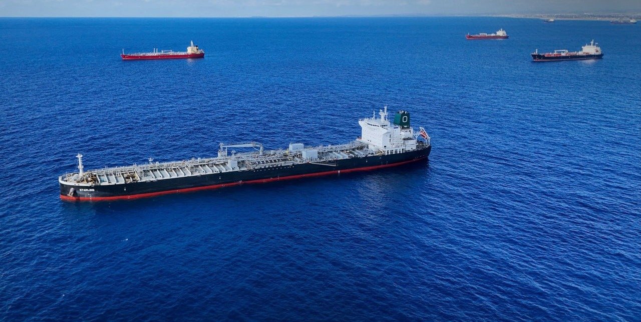 Aerial view of a large black and red oil tanker at anchor in deep blue sea, with three other ships in the distance