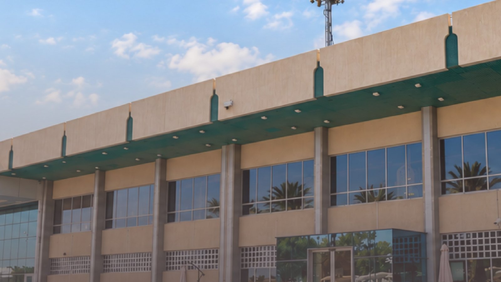 Exterior of the terminal building at Tahnoun International Airport in Al Ain, with a green canopy, glass entrance and bollards in the foreground