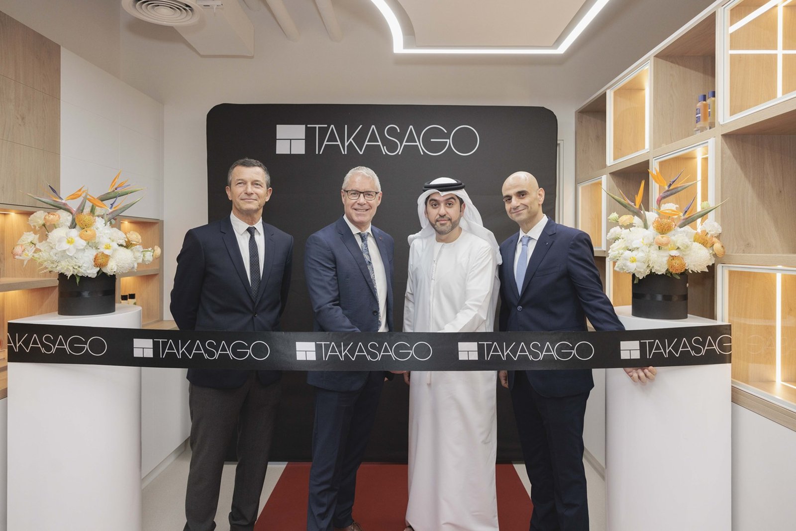 Four men in suits and traditional Emirati dress stand behind a black Takasago ribbon at the opening of a fragrance center with wooden shelving and flower displays