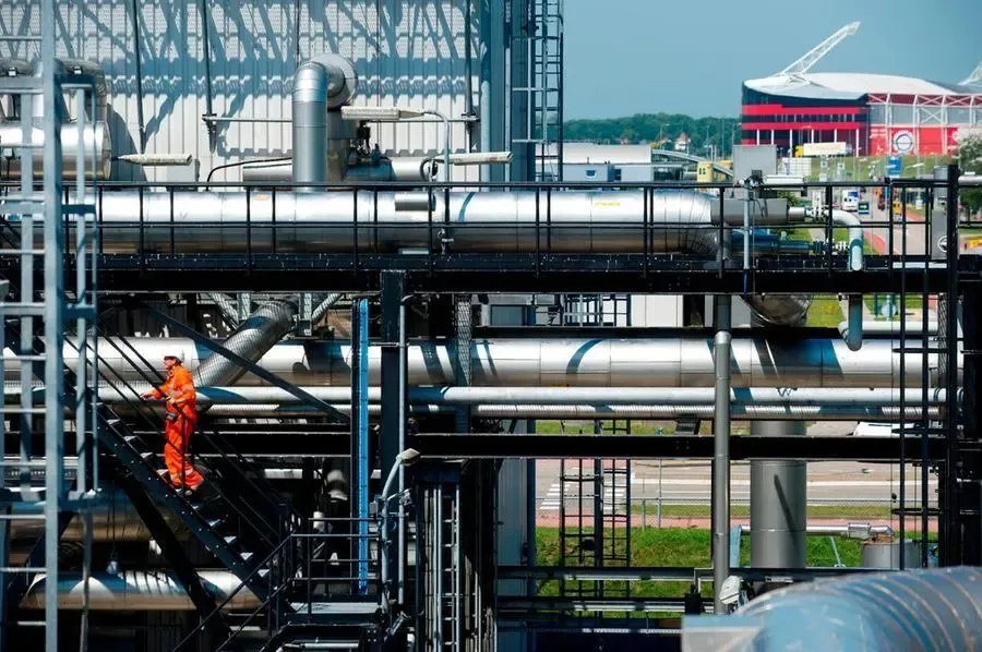 A worker in orange overalls and a hard hat climbs metal stairs among large insulated pipes at an industrial energy facility