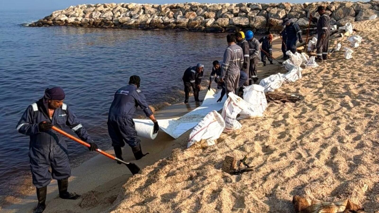 Workers in overalls laying white absorbent rolls and sandbags along the shoreline of a beach in Umm Al Quwain