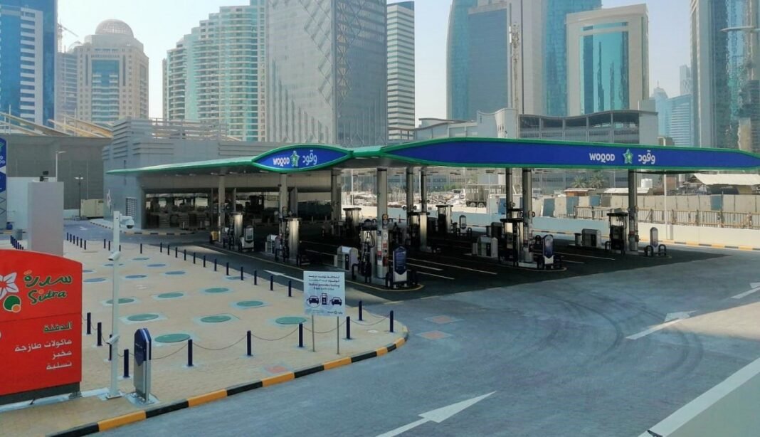 A Woqod fuel station with a blue and green canopy in front of high-rise towers in Doha
