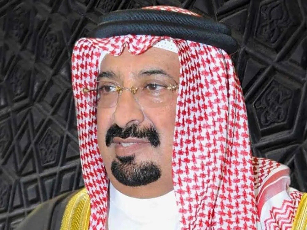 Portrait of a man wearing glasses and a red and white checked headdress in front of a dark carved wooden wall