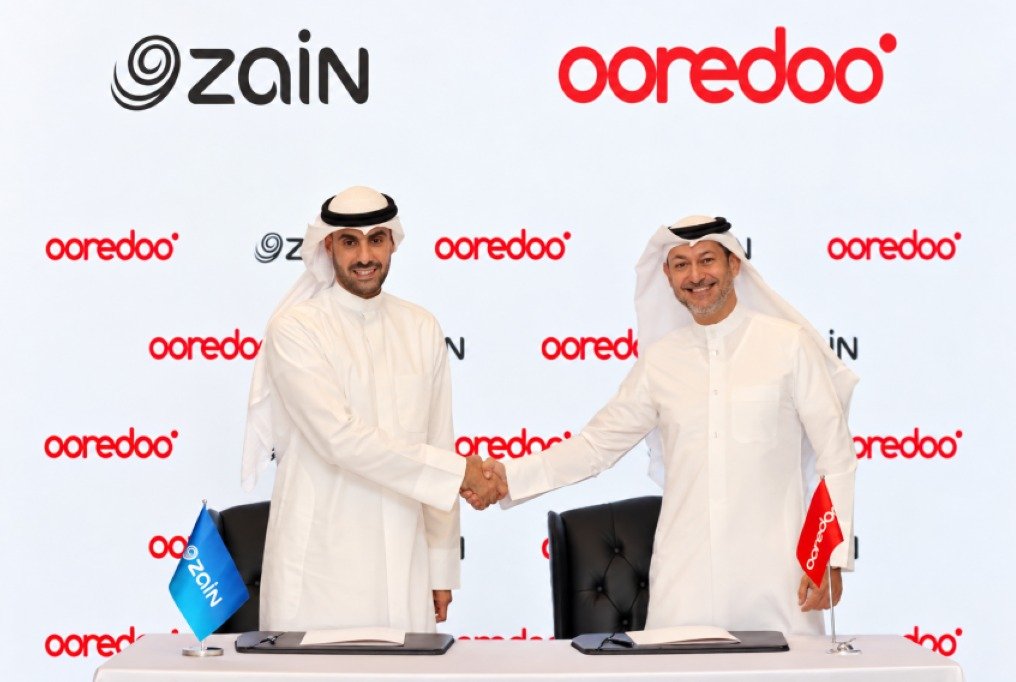 Two men in white Gulf dress shake hands in front of a backdrop printed with Zain and Ooredoo logos, with company flags on a signing table