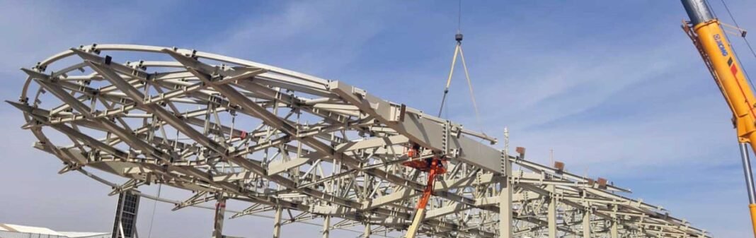 Large curved structural steel roof frame being lifted by a crane against a blue sky